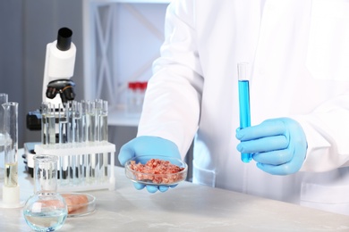 Photo of Scientist holding test tube and Petri dish with forcemeat over table in laboratory