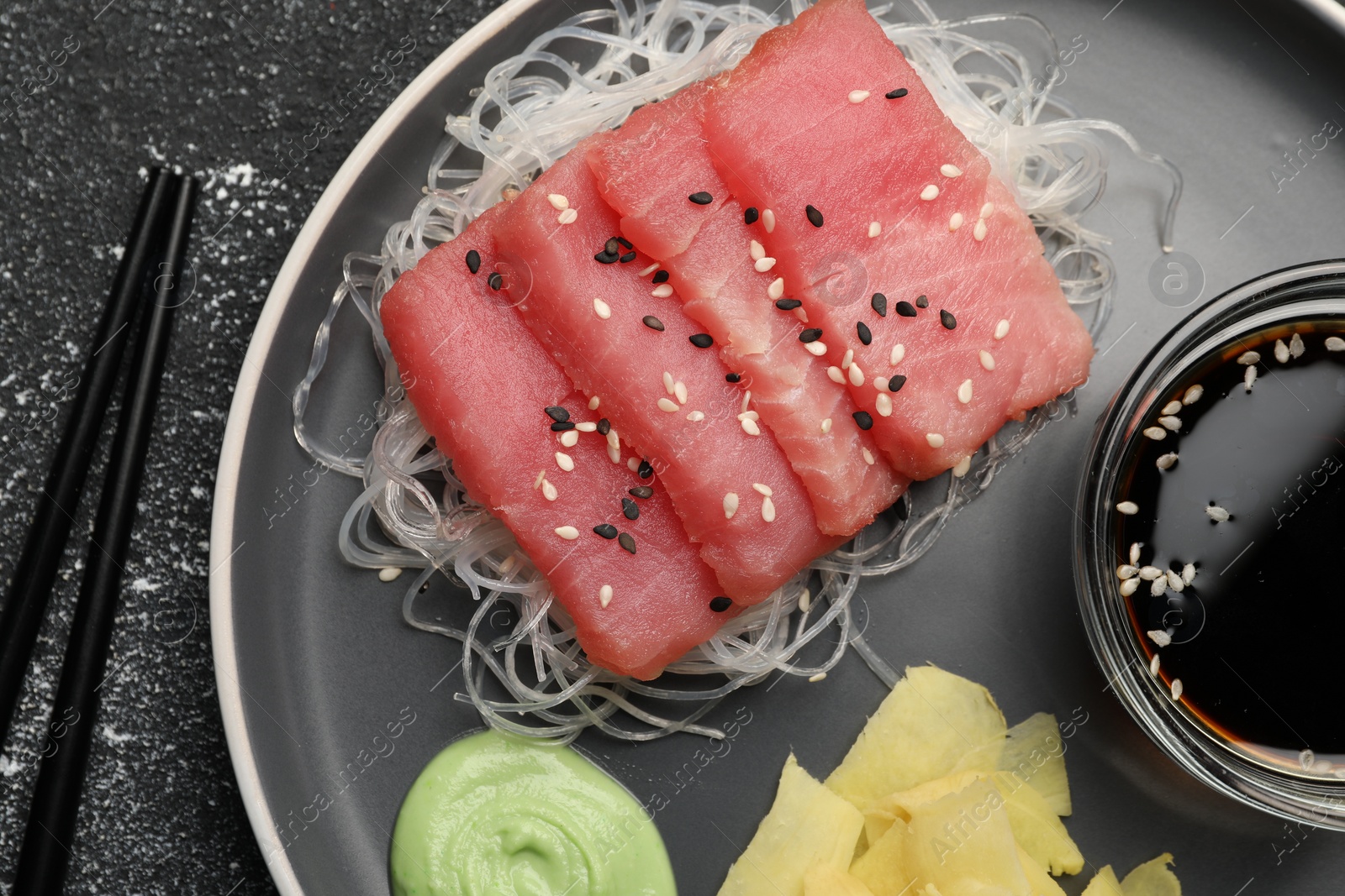 Photo of Tasty sashimi (pieces of fresh raw tuna), glass noodles, soy sauce, wasabi and ginger slices on dark table, flat lay