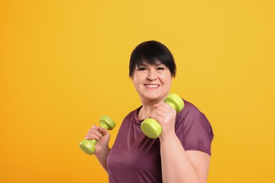 Happy overweight mature woman doing exercise with dumbbells on orange background