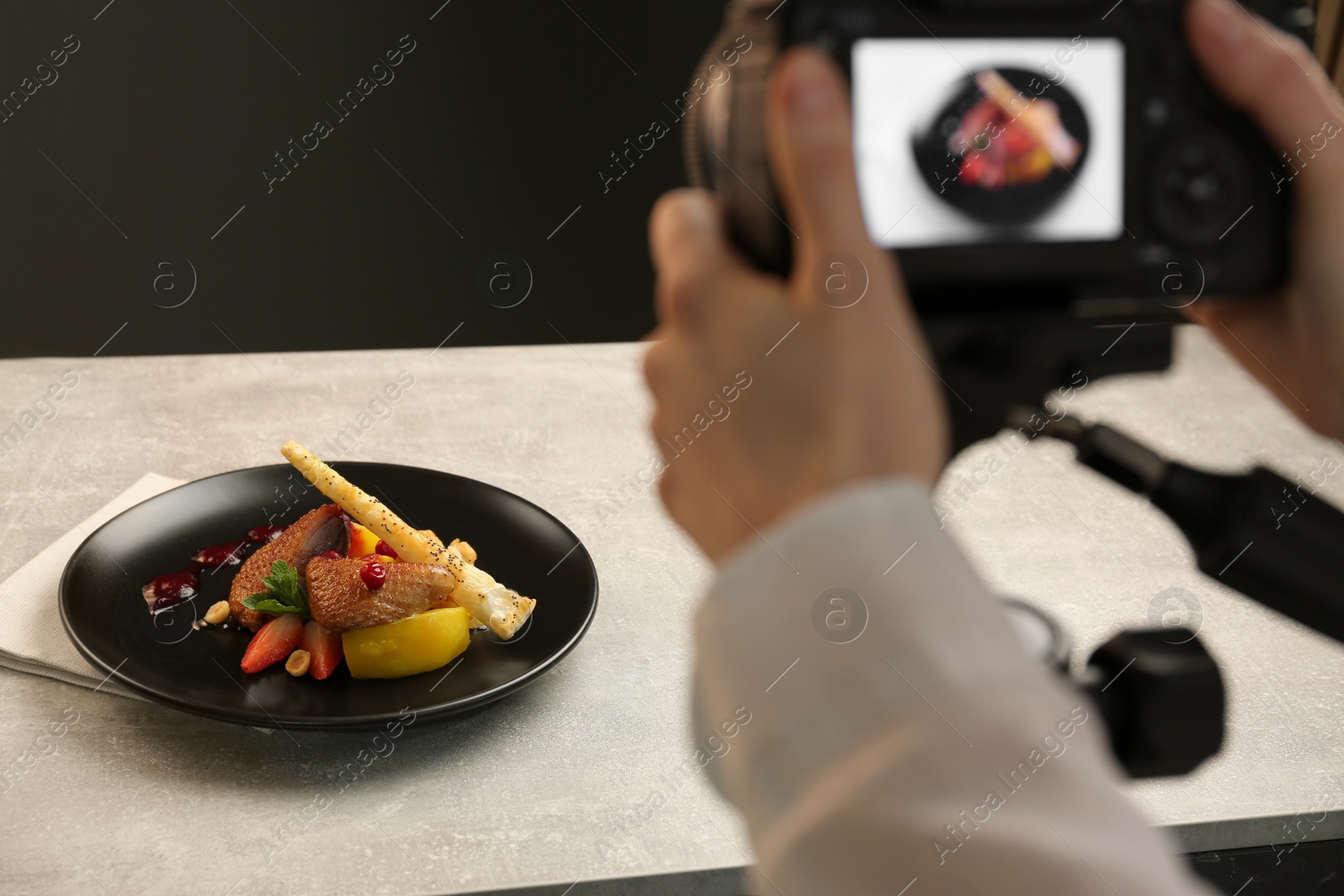 Photo of Woman taking picture of dish with chicken, parsnip and strawberries on grey table in professional photo studio, closeup. Food stylist