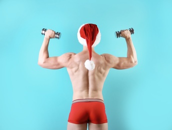 Young muscular man in Santa hat with dumbbells on color background