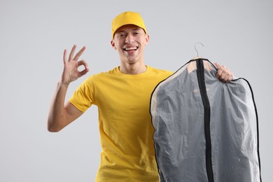 Photo of Dry-cleaning delivery. Happy courier holding garment cover with clothes and showing OK gesture on light grey background