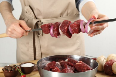 Woman stringing marinated meat and onion on skewer at table, closeup