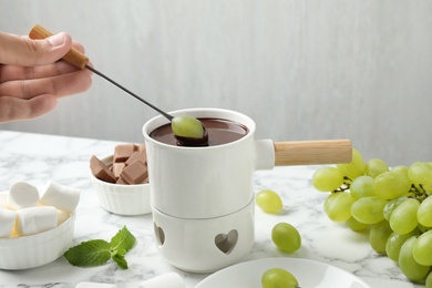 Photo of Man dipping grape into fondue pot with dark chocolate at marble table, closeup