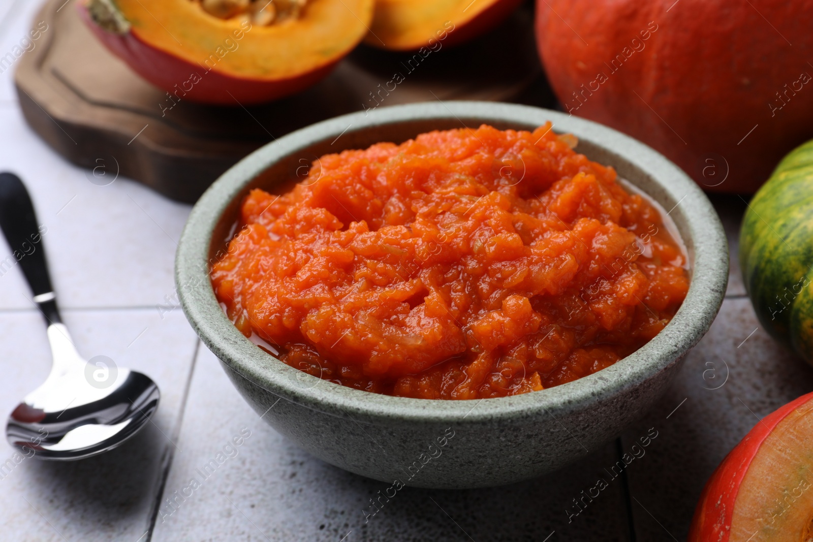 Photo of Bowl of delicious pumpkin jam and fresh pumpkin on tiled surface, closeup