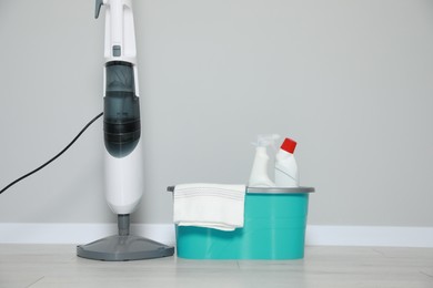 Modern steam mop and bucket with different cleaning supplies on floor near grey wall