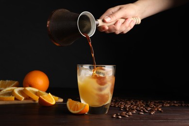 Woman pouring coffee into glass with orange juice and ice cubes at wooden table, closeup