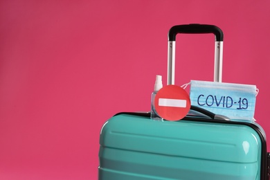 Photo of Antiseptic spray, stop sign and protective masks with inscription COVID-19 on suitcase against pink background, space for text. Travel restriction during coronavirus pandemic