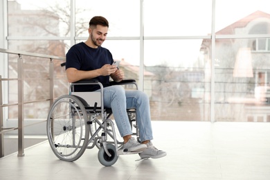 Photo of Young man in wheelchair using mobile phone near window indoors