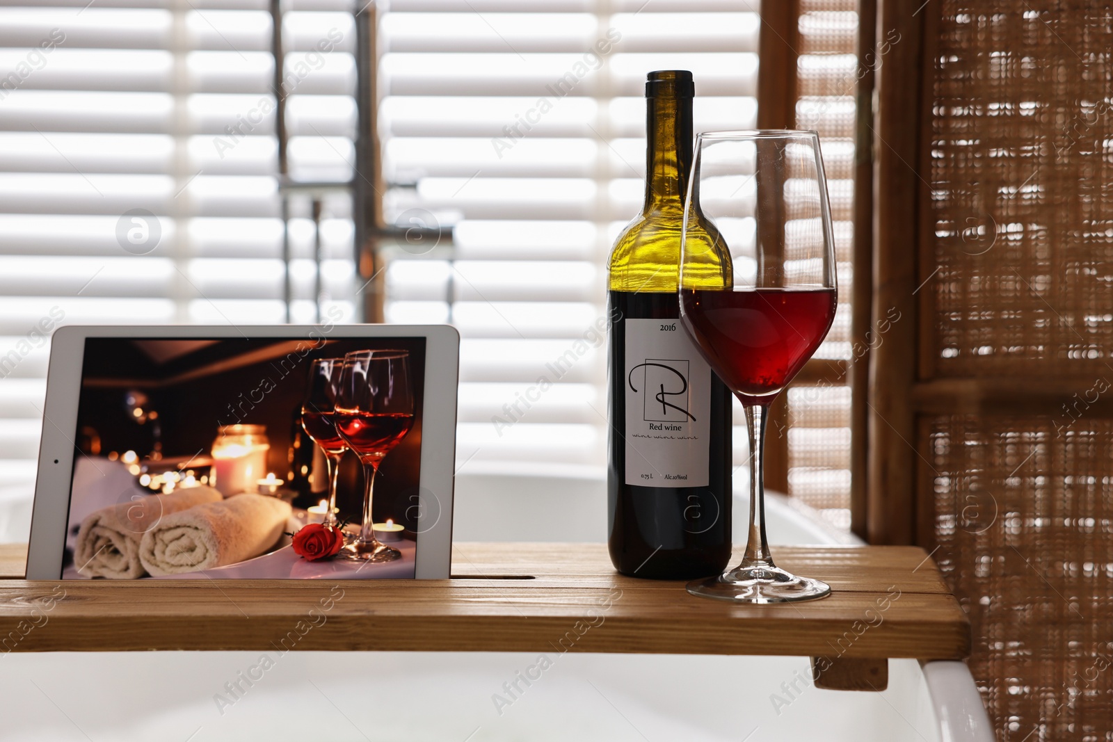 Photo of Wooden tray with tablet, glass of wine and bottle on bathtub in bathroom