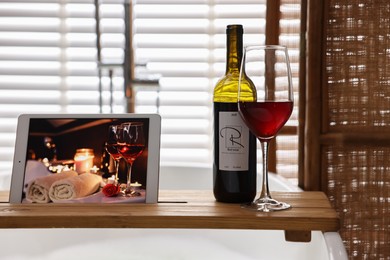Photo of Wooden tray with tablet, glass of wine and bottle on bathtub in bathroom