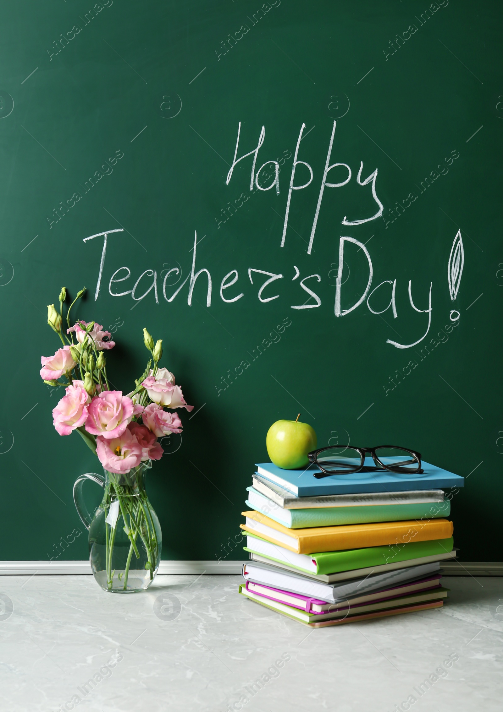 Photo of Green chalkboard with inscription HAPPY TEACHER'S DAY, vase of flowers and books on grey stone table