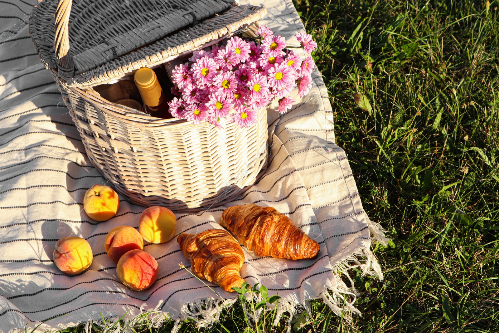 Photo of Picnic basket with flowers, bottle of wine and food on blanket outdoors