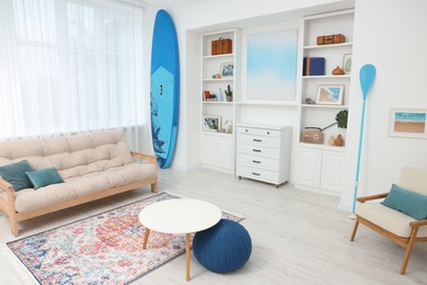 Photo of SUP board, shelving unit with different decor elements, table and pouf in room. Interior design