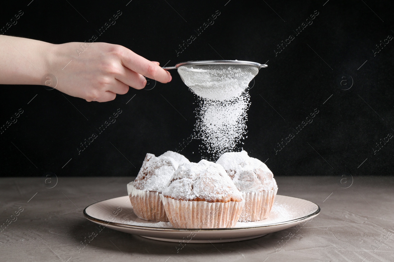 Photo of Woman with sieve sprinkling powdered sugar onto muffins at grey textured table, closeup