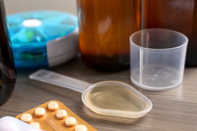 Dosing spoon with syrup, measuring cup and pills on wooden table, closeup. Cold medicine