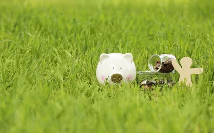 Photo of Piggy bank, wooden person and jars with coins on green grass in park. Space for text