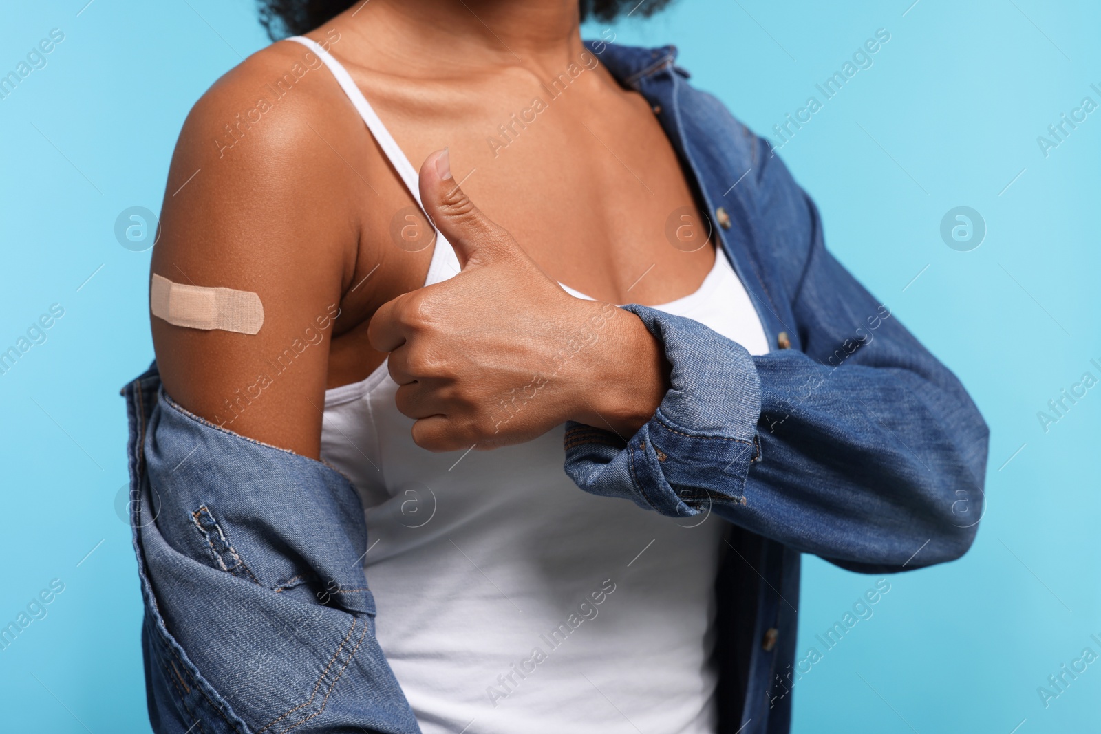 Photo of Young woman with adhesive bandage on her arm after vaccination showing thumb up against light blue background, closeup