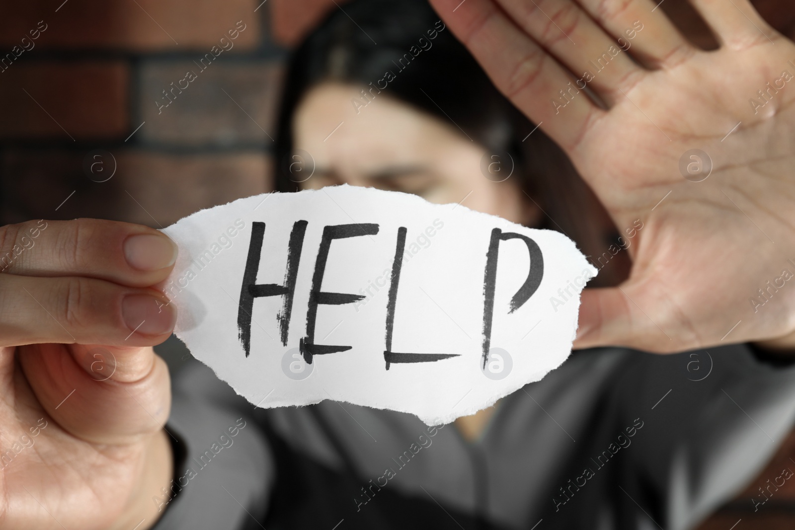 Photo of Domestic violence concept. Unhappy woman holding paper with written word Help near brick wall, focus on hands