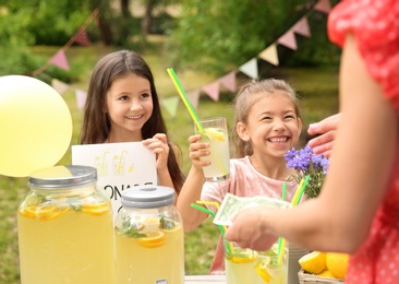 Photo of Little girls selling natural lemonade to woman at stand in park