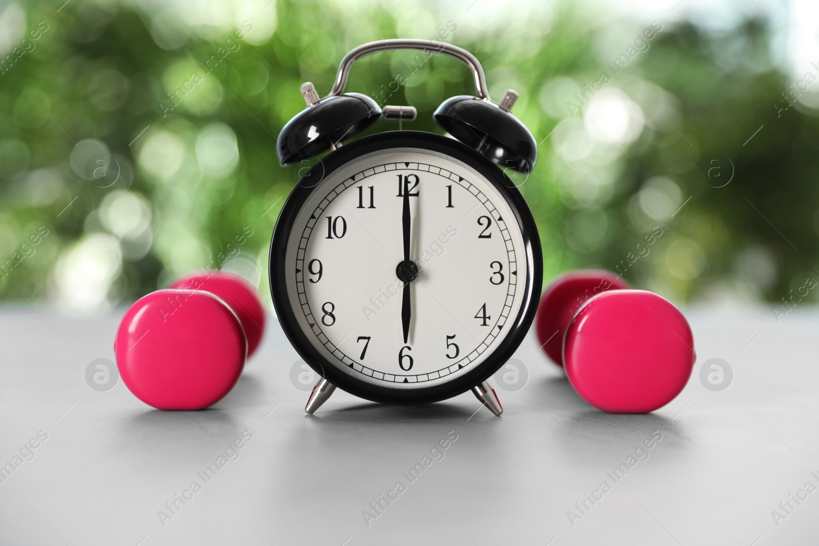 Photo of Alarm clock and dumbbells on grey table against blurred background. Morning exercise