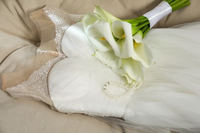 Photo of Beautiful calla lily flowers tied with ribbon, wedding dress and necklace on sofa