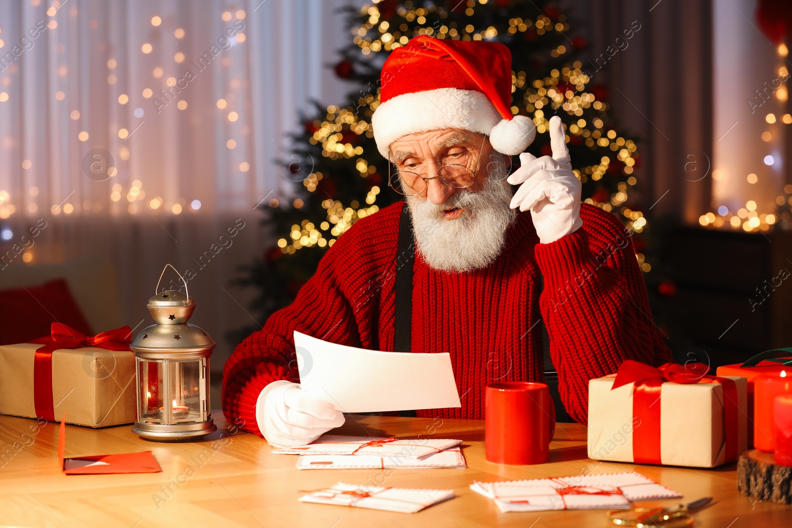 Photo of Santa Claus reading letter at his workplace in room with Christmas tree
