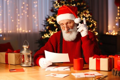 Santa Claus reading letter at his workplace in room with Christmas tree