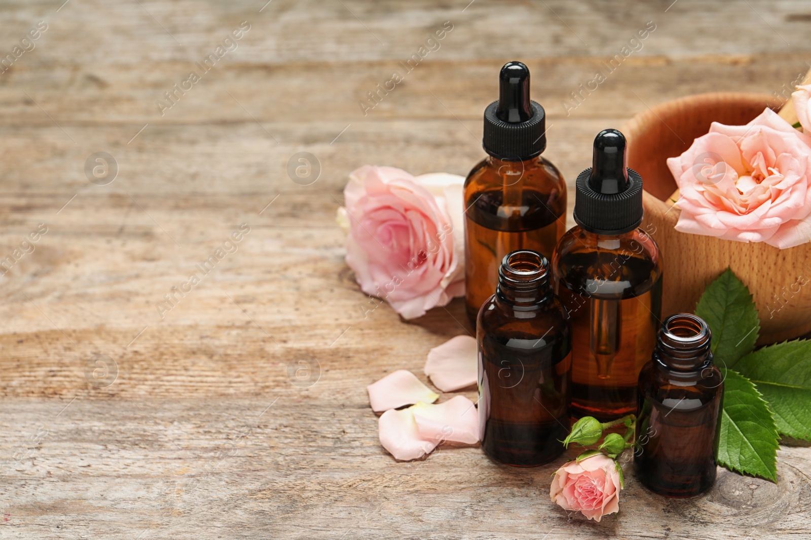 Photo of Bottles of rose essential oil and flowers on wooden table, space for text