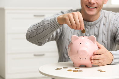 Photo of Man putting coin into piggy bank at white table indoors, closeup