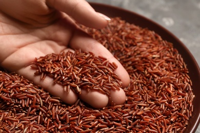 Woman holding grains near plate with brown rice on table, closeup