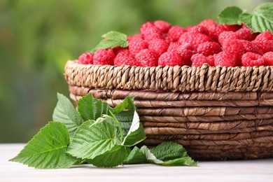 Photo of Wicker basket with tasty ripe raspberries and leaves on white wooden table against blurred green background, closeup