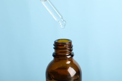 Photo of Dripping cosmetic serum from pipette into bottle on light blue background