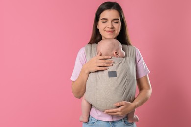 Mother holding her child in sling (baby carrier) on pink background. Space for text