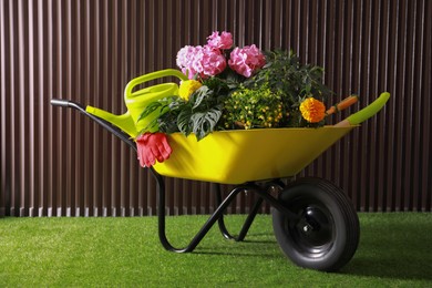 Photo of Wheelbarrow with gardening tools and plants on green grass near wood slat wall