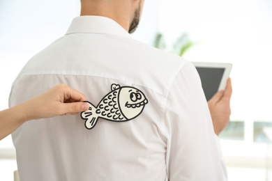 Photo of Woman sticking paper fish to colleague's back in office, closeup. April fool's day