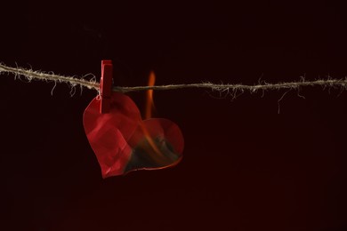 Photo of Crumpled red paper heart burning on rope against burgundy background, space for text. Broken heart