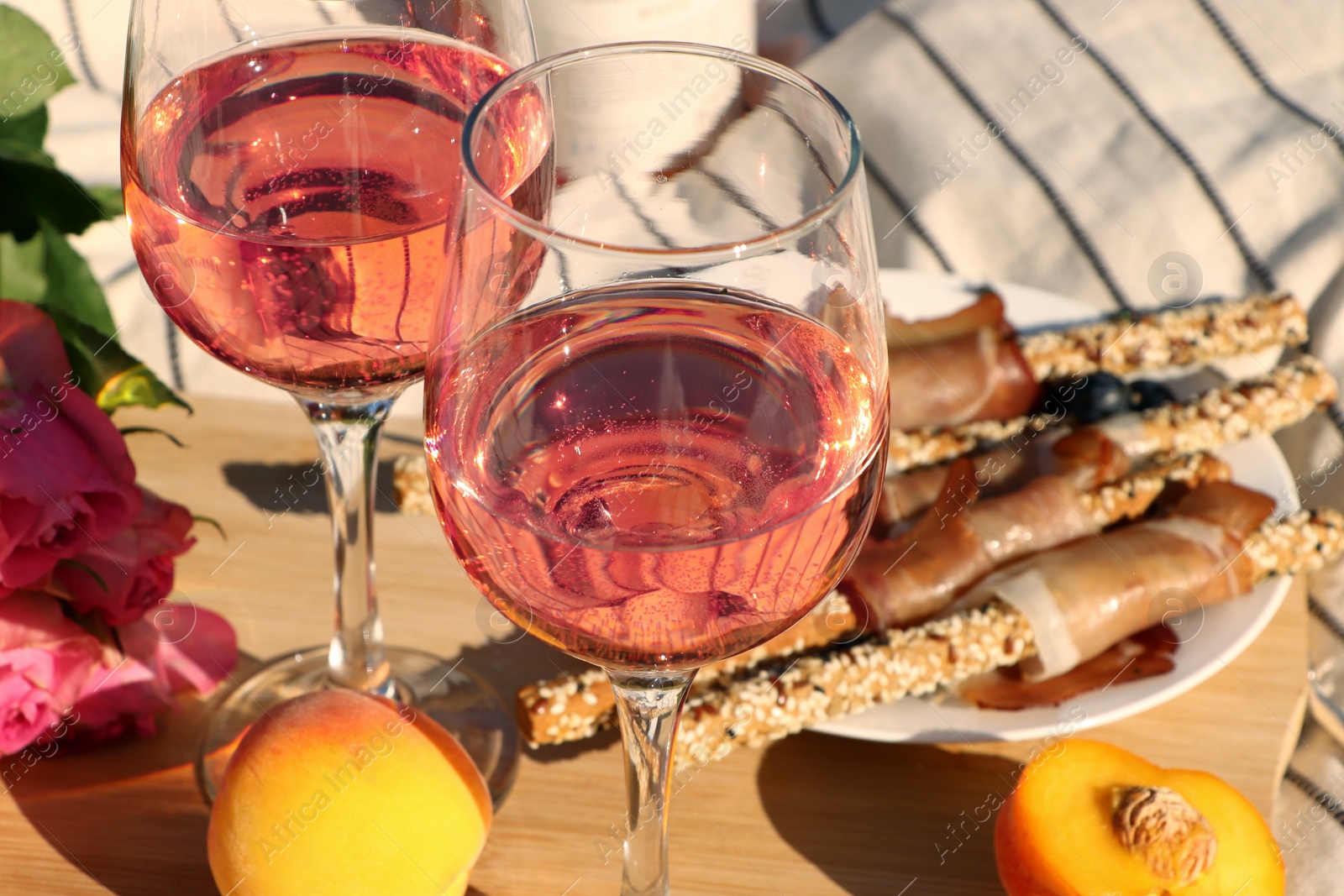 Photo of Glasses of delicious rose wine, flowers and food on white picnic blanket, closeup