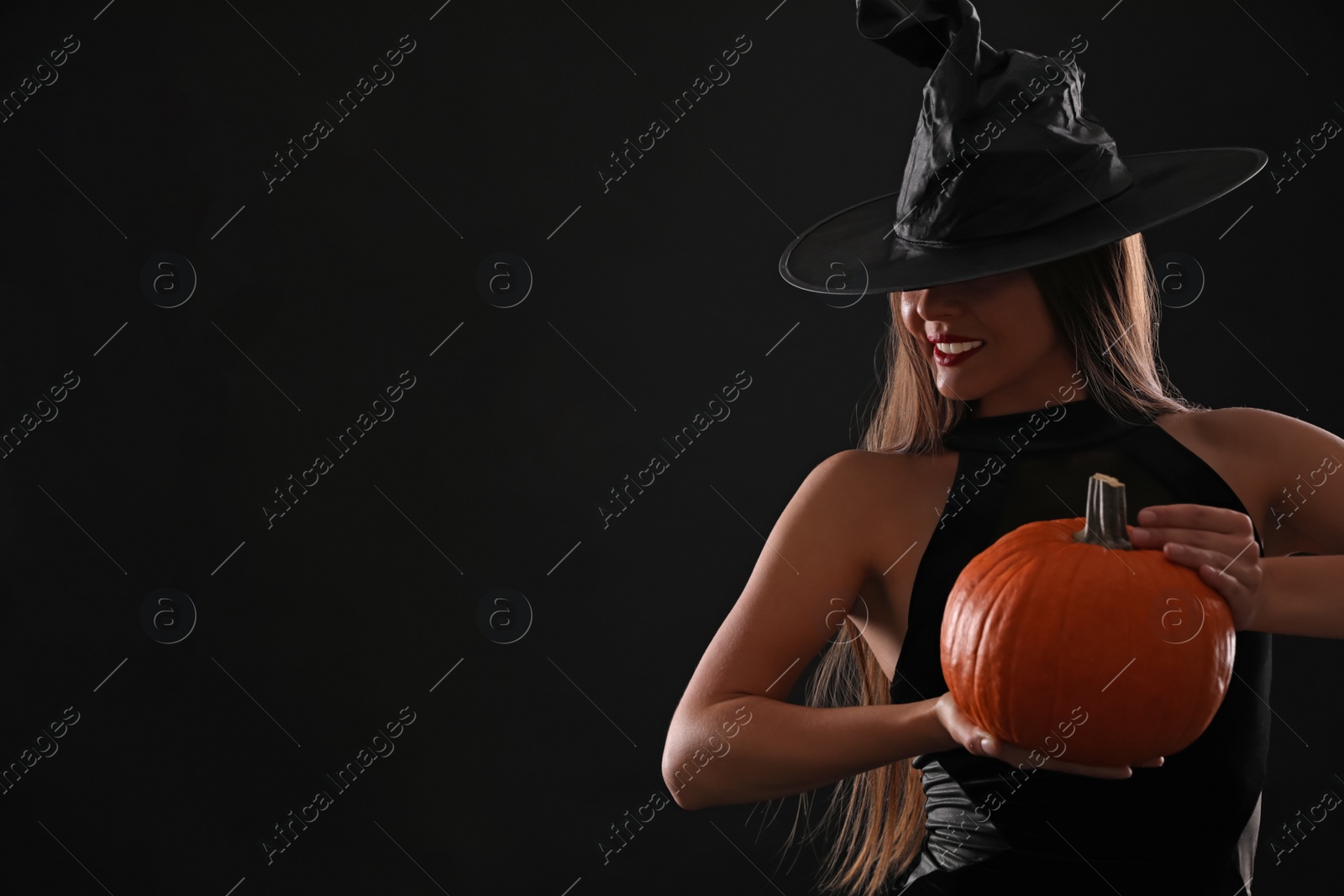 Photo of Young woman wearing witch costume with pumpkin on black background, space for text. Halloween party