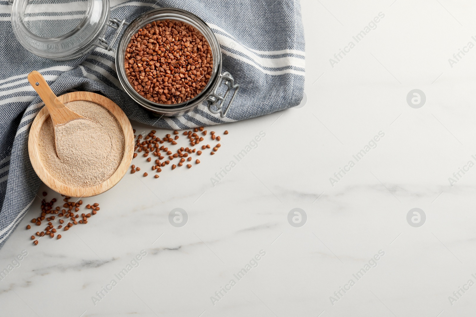 Photo of Bowl of buckwheat flour and glass jar with grains on white marble table, flat lay. Space for text