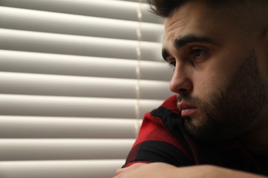 Photo of Sad young man near closed blinds at home. Space for text