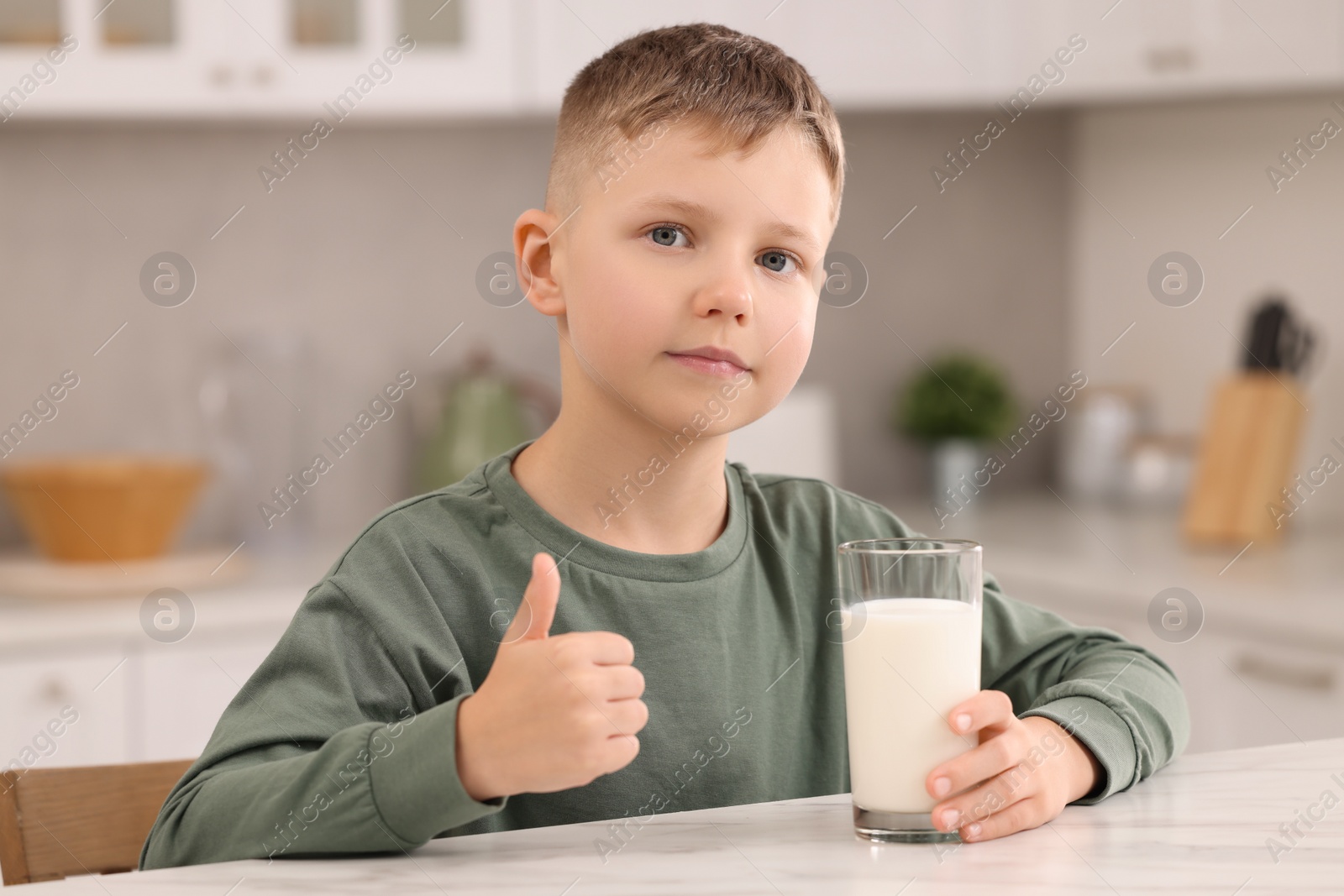 Photo of Cute boy with glass of fresh milk showing thumb up at white table in kitchen
