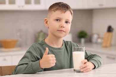 Cute boy with glass of fresh milk showing thumb up at white table in kitchen