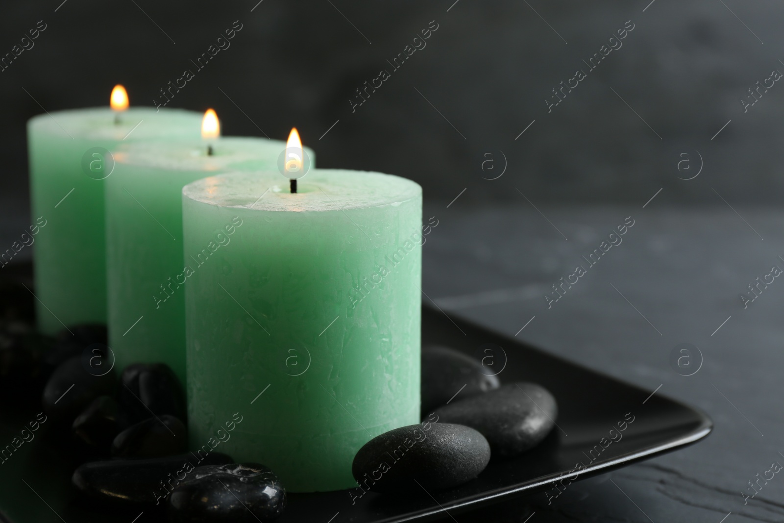 Photo of Dark plate with three burning candles and rocks on table, space for text