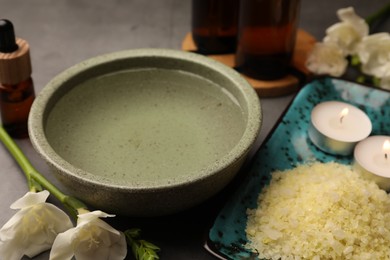 Photo of Bowl of essential oil, sea salt and beautiful flowers on grey table, closeup. Aromatherapy treatment