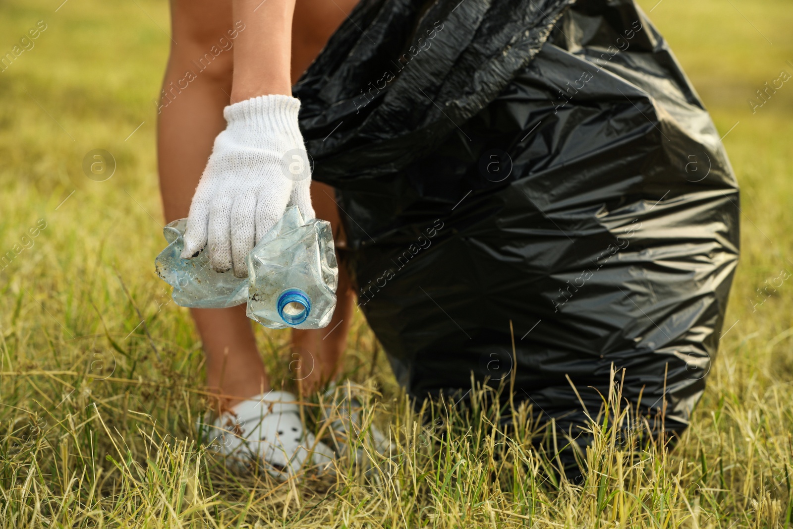 Photo of Woman in gloves with trash bag picking crumpled bottle from grass outdoors, closeup