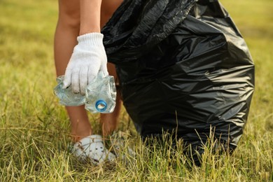 Photo of Woman in gloves with trash bag picking crumpled bottle from grass outdoors, closeup