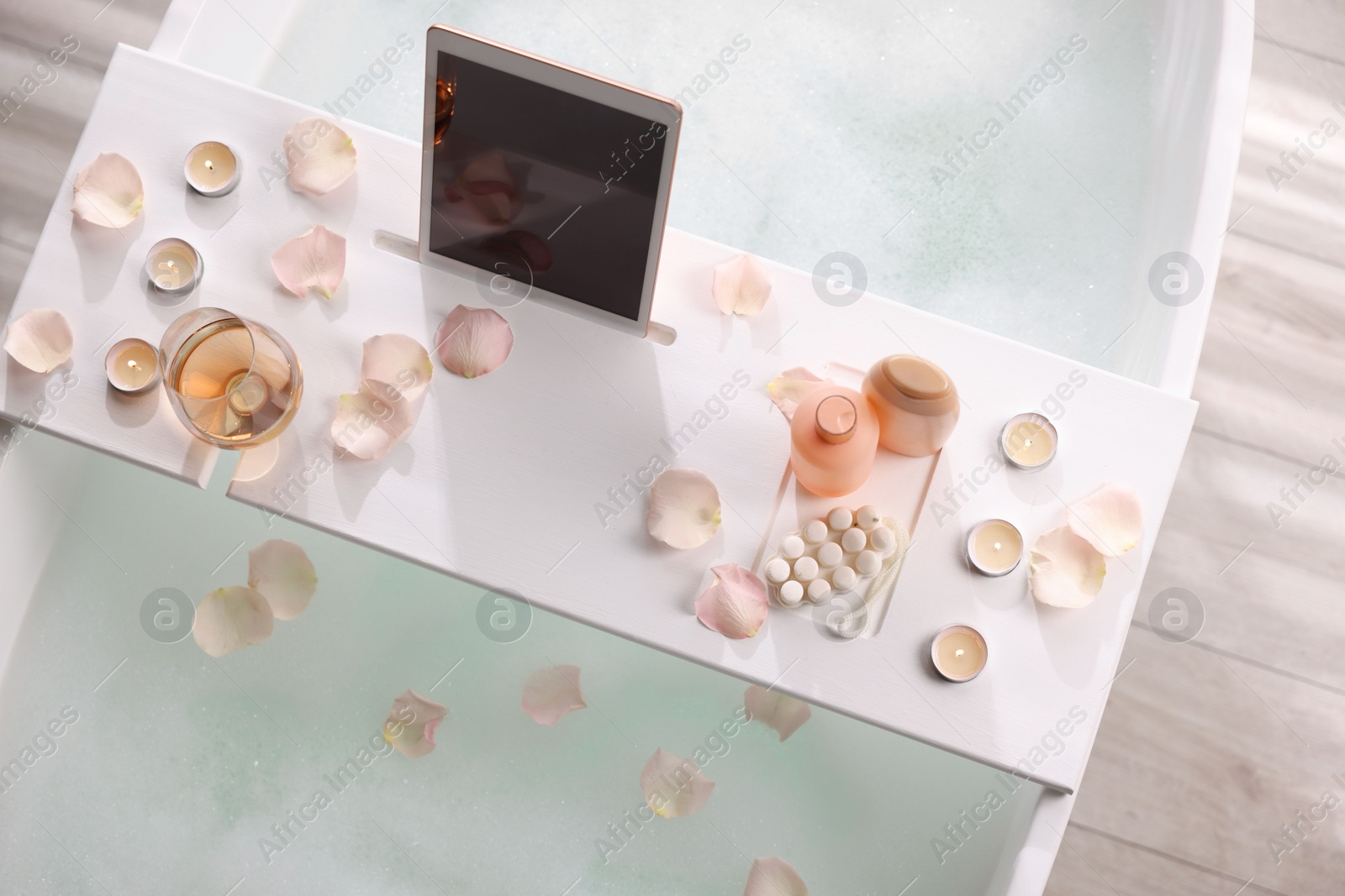 Photo of Wooden tray with tablet, wine, toiletries and flower petals on bathtub in bathroom, above view