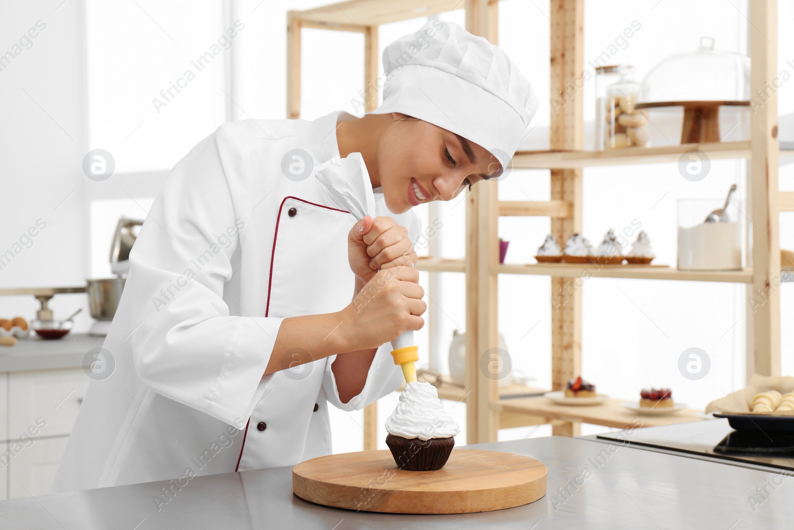 Photo of Young female pastry chef decorating cupcake with cream at table in kitchen