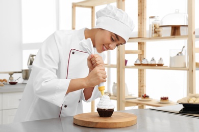 Photo of Young female pastry chef decorating cupcake with cream at table in kitchen